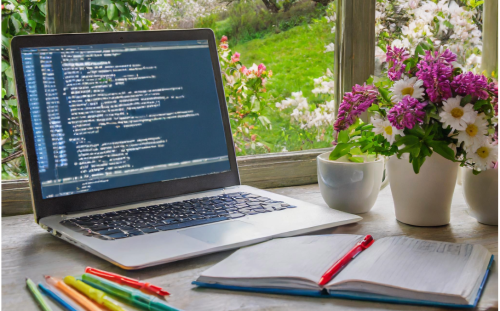 View of a desktop with laptop displaying HTML code. Open notebook with pens and pencils and a vase of blooming spring flowers. Flowers and a green hillside are also visible out the window.